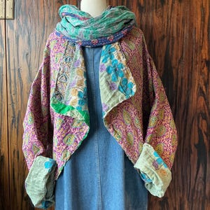 May include: A long-sleeved, open-front jacket featuring a vibrant purple and gold paisley pattern, layered over a blue denim dress. A matching green and blue floral patterned scarf is wrapped around the neck, with wide cuffs folded back to reveal the inner lining.