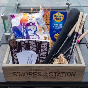 May include: A wooden S'mores Station box filled with marshmallows, graham crackers, Hershey's chocolate bars, and roasting sticks. The box has the text "Screer Family S'mores Station" engraved on the front. A fire pit with a glass enclosure is in the background.
