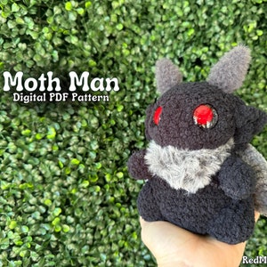 May include: A black and gray plush toy with red eyes, resembling a moth. The toy is being held in a hand. The text "Moth Man Digital PDF Pattern" is visible in the image.