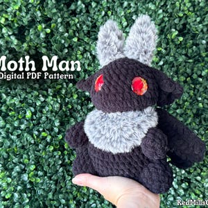 May include: Black crocheted moth plush toy with red eyes and gray fur accents. The toy is being held by a hand. The text "Moth Man Digital PDF Pattern" is visible in the image.