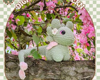 Puede incluir: Un juguete de axolotl dragón amigurumi tejido a crochet con un cuerpo verde, una cola rosa y una flor rosa en la cabeza. El juguete está sentado en una rama de árbol con flores rosas en el fondo. El texto "Dahlia the Axolotl Dragon" está escrito debajo del juguete.