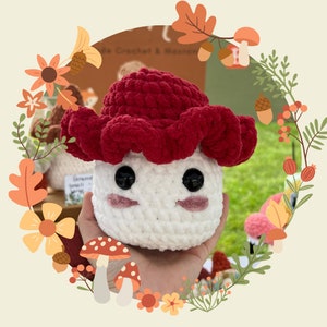May include: A crocheted mushroom amigurumi toy with a red cap and white body. The toy has a cute face with black eyes and pink cheeks. The toy is being held by a hand.