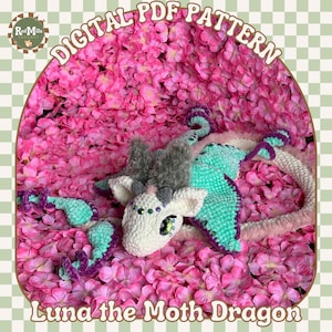 Peut inclure: Un jouet en crochet représentant un dragon papillon avec un corps blanc, des ailes vertes et des accents roses et violets. Le jouet est posé sur un lit de pétales de fleurs roses. Le texte "Luna the Moth Dragon" est écrit sous le jouet.