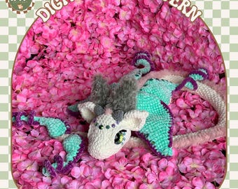 Puede incluir: Un juguete de dragón de polilla de crochet con un cuerpo blanco, alas verdes y detalles en rosa y morado. El juguete está acostado sobre un lecho de pétalos de flores rosas. El texto "Luna the Moth Dragon" está escrito debajo del juguete.