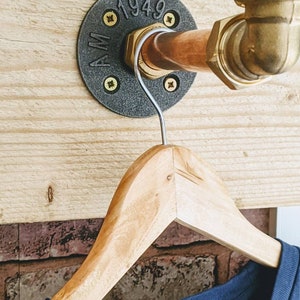 May include: A copper pipe coat rack with a black metal flange. The flange has the text "AM 1949" on it. A wooden hanger is hanging from the pipe.