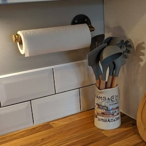 May include: A kitchen setting with a white paper towel roll on a black and brass holder. A ceramic jar with kitchen utensils and a wooden worktop are also in view.