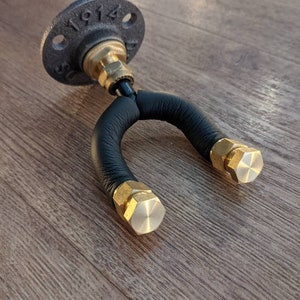 Może przedstawiać: Czarny skórzany hak na gitarę ze złotymi metalowymi akcentami i czarną żeliwną flanszą rury z wytłoczonym numerem "1914".