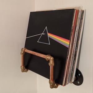 May include: A copper pipe record shelf with gold fittings holds a stack of vinyl records, including one with the iconic "Dark Side of the Moon" album cover art featuring a prism and a rainbow.