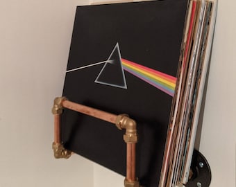 May include: A copper pipe record shelf with gold fittings holds a stack of vinyl records, including one with the iconic "Dark Side of the Moon" album cover art featuring a prism and a rainbow.