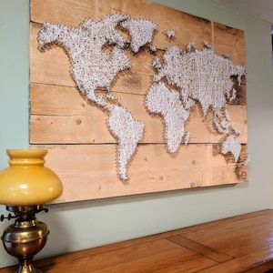 May include: A wooden world map created with nails and white string. The continents are outlined with string on a light brown wooden background. A vintage brass and yellow lamp sits on a wooden table in the foreground.