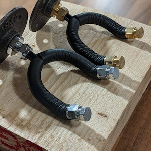 Könnte beinhalten: Zwei schwarze Metallrohr-Kleiderhaken mit geschwungenem Design. Die Haken sind auf einer Holzplanke montiert. Die Haken haben Messing- und silberfarbene Beschläge. Die Haken sind für die Wandmontage konzipiert.