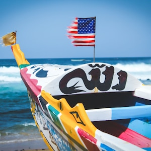 Op de afbeelding: Een kleurrijk houten boot met een zwart-wit ontwerp op de boeg. De boot ligt aangemeerd aan een zandstrand met blauwe oceaan golven op de achtergrond. Een Amerikaanse vlag en een gele vlag wapperen vanaf de boot.