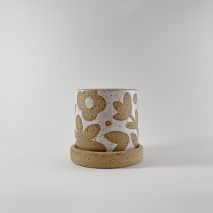 May include: A white ceramic planter with a brown floral design. The planter has a matching saucer.