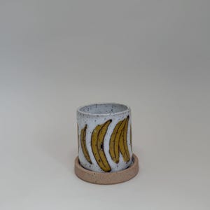 May include: A cylindrical ceramic planter with a speckled white finish. The planter features a repeating pattern of yellow bananas. It sits on a small, round, unglazed, tan-colored saucer. The design is simple and modern.