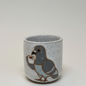 Puede incluir: Una taza de cerámica cilíndrica con un esmalte blanco moteado. El exterior presenta una ilustración pintada a mano de una paloma gris que sostiene una pequeña taza blanca. La taza tiene un borde redondeado suave y una base resistente y funcional.