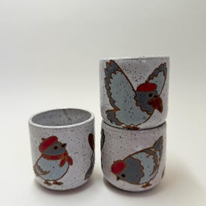 Puede incluir: Tres tazas de cerámica con esmalte blanco moteado, cada una con ilustraciones pintadas a mano de palomas con boinas y bufandas rojas. Las palomas se muestran en varias posturas, incluyendo de pie y volando, con alas grises y detalles rojos.