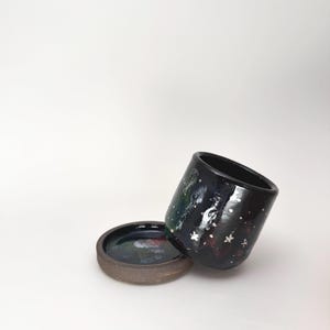 May include: A black ceramic planter with a glossy finish featuring a galaxy-inspired pattern of white dots and star shapes. The cylindrical pot rests at an angle against its matching circular saucer, which has a dark, mottled interior surface.