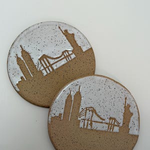 May include: Two round ceramic coasters featuring a speckled tan clay base and a white glazed upper section. The design depicts a New York City skyline silhouette including the Statue of Liberty, the Brooklyn Bridge, and skyscrapers in brown.