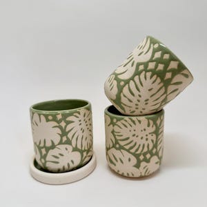 May include: Two cylindrical ceramic planters with a matte sage green and off-white monstera leaf pattern. One planter sits upright on a matching circular off-white saucer, while the second planter is tilted and balanced on top of a third planter.