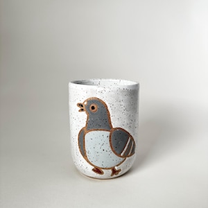 May include: A ceramic mug with a hand-painted pigeon design. The mug is white with black speckles, and the pigeon is in shades of grey, blue and brown. The mug has a rounded shape and a speckled texture. The design is simple and cartoonish.
