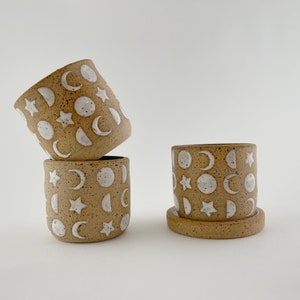 May include: Four small, speckled ceramic pots with white moon and star designs. One pot sits on a matching saucer. The other three pots are stacked, showcasing the celestial patterns. The pots have a natural, earthy tone.