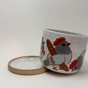 May include: A speckled white ceramic planter with a matching saucer, featuring hand-painted illustrations of grey birds wearing red berets. The cylindrical pot has a smooth interior and a textured exterior finish with brown speckles throughout.