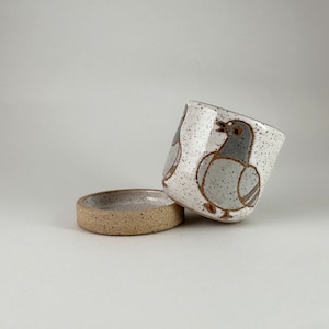 May include: A ceramic cup and saucer set. The cup is white with a speckled pattern and a brown outline of a pigeon. The saucer is a speckled tan colour. The cup is tilted, and the saucer is on the ground.