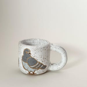 May include: A speckled ceramic mug with a gray pigeon illustration. The mug has a rounded handle and a white speckled finish. The pigeon design features gray and gold accents.