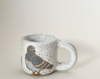 May include: A speckled ceramic mug with a gray pigeon illustration. The mug has a rounded handle and a white speckled finish. The pigeon design features gray and gold accents.