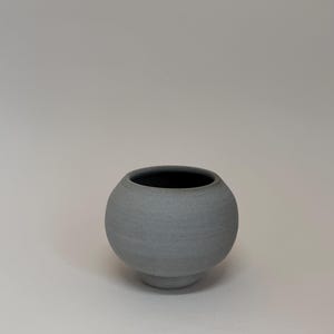 May include: A small, gray ceramic vase with a rounded body and a dark interior. The vase sits on a short, circular base. The surface has a textured appearance, and the overall design is minimalist and modern.