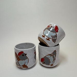 May include: Three white ceramic cups with a speckled finish, each featuring hand-painted illustrations of grey pigeons wearing red berets. One cup is balanced on top of another, while the third sits beside them, showcasing the detailed bird art.