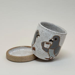 May include: A speckled white ceramic mug with a matching saucer. The mug features hand-drawn illustrations of grey pigeons with brown accents. The saucer is a flat, round dish with a raised edge. The mug is tilted, showing the interior.