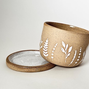 May include: A speckled, tan ceramic planter with a matching saucer. The planter features white floral designs around the exterior. The saucer is light gray with a brown rim. The planter is slightly tilted, revealing its interior.