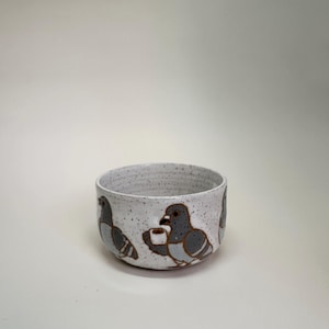 May include: A speckled ceramic bowl with a rounded shape. The bowl features a repeating pattern of gray and brown pigeon illustrations. The pigeons are holding small cups. The bowl has a white interior and exterior.