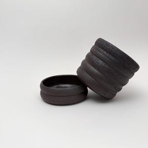 Könnte beinhalten: Eine dunkel anthrazitfarbene Keramiktasse und eine passende flache Schale. Beide Teile haben eine raue, texturierte Außenfläche mit horizontalen Rillen und eine glatte, dunkle Innenfläche. Die Tasse ist gegen die Seite der Schale gelehnt.