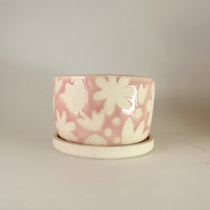 May include: A ceramic planter with a matching saucer. The planter is light pink with a white floral pattern. The saucer and the planter have a speckled texture. The planter is designed for indoor or outdoor use.
