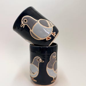 May include: Two cylindrical ceramic cups with a glossy dark navy blue glaze. The cups feature hand-painted illustrations of grey pigeons with light grey wings and orange feet, outlined in a thin, light-colored line against the dark background.