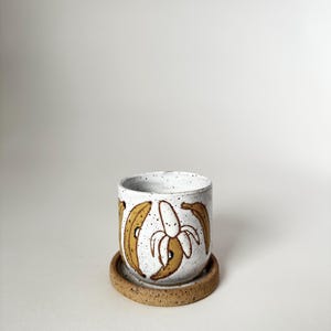 May include: A speckled white ceramic mug with a brown banana design and a matching saucer. The mug features a peeled banana in the center and whole bananas around the sides. The mug and saucer are on a white background.