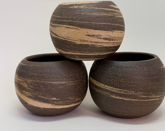 Pode incluir: Três pequenos vasos redondos de cerâmica em um arranjo empilhado. Os vasos são marrons escuros com padrões em espiral horizontais marrons mais claros. As aberturas dos vasos são visíveis, revelando um interior escuro. O fundo é branco liso.