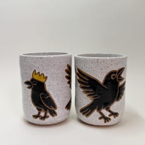 May include: Two cylindrical ceramic tumblers with a white speckled glaze. The left tumbler features a black bird wearing a yellow crown. The right tumbler displays a black bird with wings spread wide. Both vessels have a smooth, glossy finish.