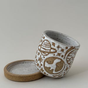 May include: A white ceramic mug with a brown speckled rim and base, featuring a hand-drawn space-themed design. The design includes planets, stars, and the Earth. A matching saucer is next to the mug.