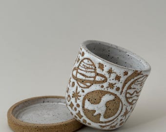 May include: A white ceramic mug with a brown speckled rim and base, featuring a hand-drawn space-themed design. The design includes planets, stars, and the Earth. A matching saucer is next to the mug.