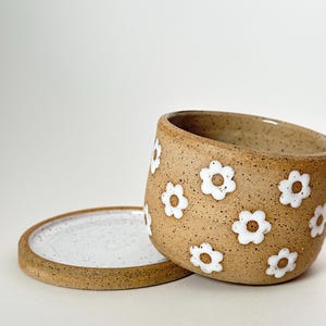 Puede incluir: Una maceta de cerámica moteada de color marrón claro con una tapa a juego. La maceta está decorada con diseños blancos en forma de margarita. La tapa es redonda y plana, con una superficie blanca moteada. La maceta está ligeramente inclinada.