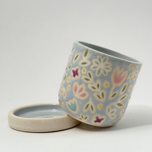 May include: A light blue ceramic planter with a matching circular saucer. The exterior of the planter features a carved, repeating pattern of stylized flowers and leaves in shades of pink, purple, yellow, and green, with a smooth glazed finish.