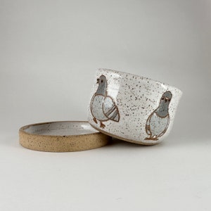 以下が含まれることがあります： 2匹の灰色のハトのイラストと、お揃いの受け皿が付いた、斑点のある白いセラミックボウル。ボウルはわずかに傾斜したデザインです。受け皿は明るい茶色です。