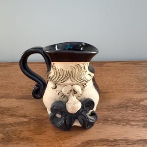 Peut inclure: Une tasse en céramique avec un visage. La tasse a une anse et un bord noirs, avec un visage beige, un nez blanc et une moustache noire. La tasse est posée sur une surface en bois.