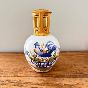 May include: A white ceramic fragrance lamp with a rounded body featuring a hand-painted blue rooster motif, green foliage, and yellow accents. The top of the vessel is fitted with a decorative, perforated gold-toned metal crown cap.