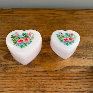 May include: Two white heart-shaped trinket boxes with floral designs on the lids. The flowers are pink, red, and blue, with green leaves. The boxes are on a wooden surface.