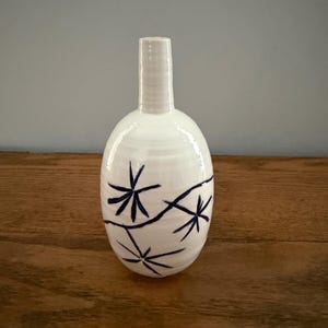 May include: A white ceramic vase with a narrow neck and an oval body. The surface features a hand-painted dark blue pattern consisting of three star-like floral motifs connected by thin, curved lines that wrap around the rounded exterior.