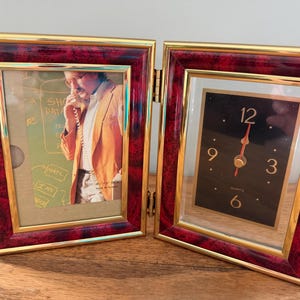 May include: A hinged, dual-frame display. One side features a photo of a person on the phone, the other a black quartz clock with gold hands and numerals. The frames have a gold trim and a burgundy and gold patterned border.